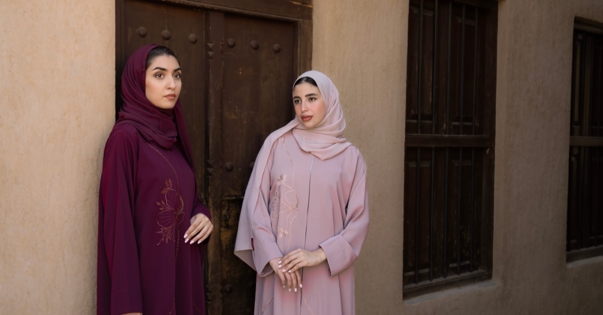 Two women wearing azael's abayas standing against a textured wall with a door.